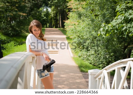 Smiling long haired young woman in white tunic and cutoffs, leaning against a white plank bridge railing and looking downwards, stilettos in hand, trees and paved road in background