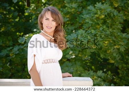 Smiling long haired young woman in white tunic and pearls necklace leaning on balustrade, looking at camera, trees in background