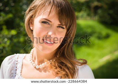 Smiling long haired young woman in white tunic and pearls necklace, looking at camera, trees in blurry background