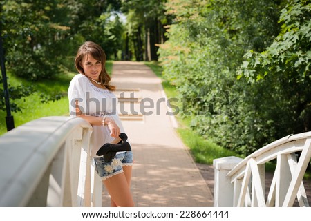 Smiling long haired young woman in white tunic and cutoffs, leaning against a white plank bridge railing and looking frowningly at camera, stilettos in hand, trees and paved road in background