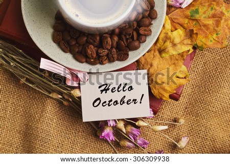 Tag with words hello october and the cup of coffee lying on the books with dry yellow maple leaves and everlasting flowers on sackcloth background