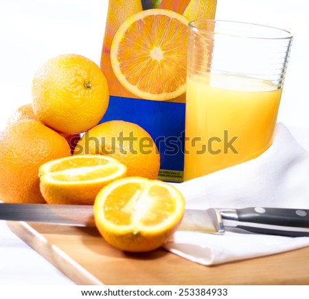 Freshly squeezed orange juice in highball tumbler glass on chopping block with whole and half sliced oranges, knife, carton and hand towel isolated