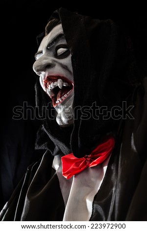 Hooded Vampire puppet with white eyes, fangs, red box tie and smartly dressed  is sneering. Horror photo celebrating Halloween, black background, isolated. Empty space for text and typography.