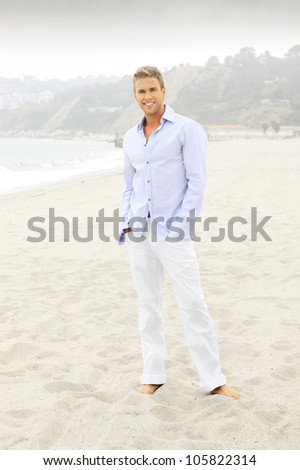 Relaxed smiling man in white pants and elegant blue shirt and bare feet on the beach