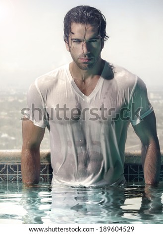 Gorgeous male model in soaked wet t-shirt standing in luxurious swimming pool with city background
