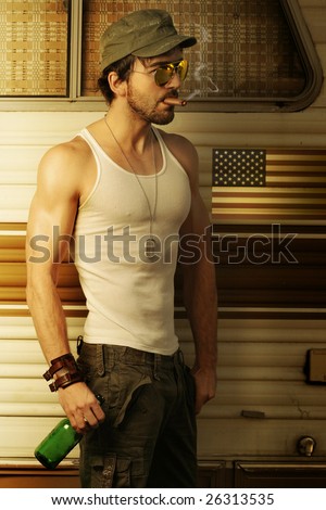 Young sexy man in white tank top and army hat smoking cigar and holding beer in front of retro trailer