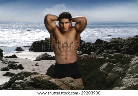 Shirtless bodybuilder flexing muscles on the beach with blue sky and ocean