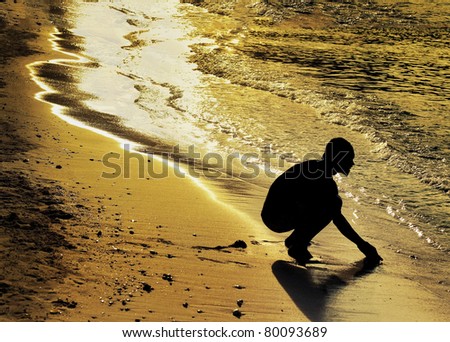 A silhouette male figure bending down touching ocean water at coastline in golden sunset light