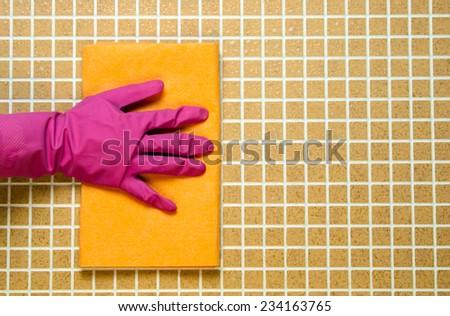 One hand in pink rubber gloves with yellow cloth on a background tile