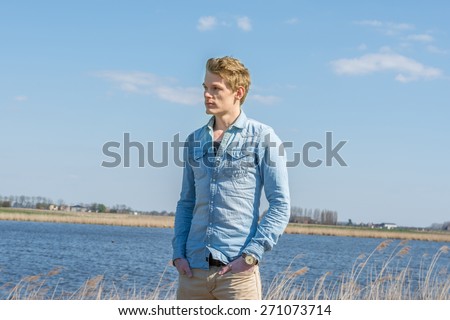 Young handsome man standing at water. Wearing blue blouse and brown pants