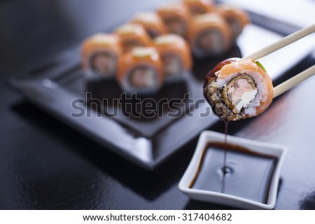 Sushi roll with cream cheese and fried salmon. Topped with raw salmon and lime. Being picked up with chopsticks from soy sauce, with more rolls on black dish in the background. Soy sauce dripping.