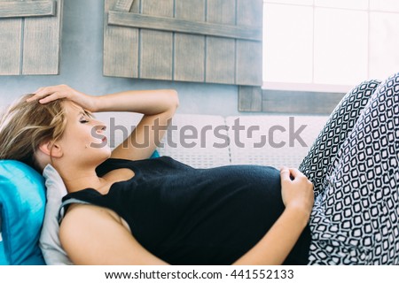 Young pregnant woman having a headache and she is lying on the sofa holding her belly and forehead