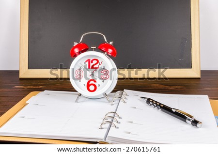 Alarm clock with notebook and black pen on wooden board and blackboard background