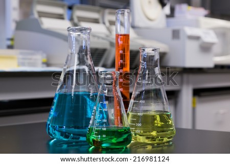 Different color liquids in flasks and a measuring cylinder with a laboratory background.
