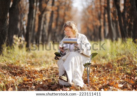 Young happy woman in the autumn sitting on a chair, wrapped in a blanket against the cold. poured from a thermos of hot tea, drink it. Peaceful life. Trees with falling leaves