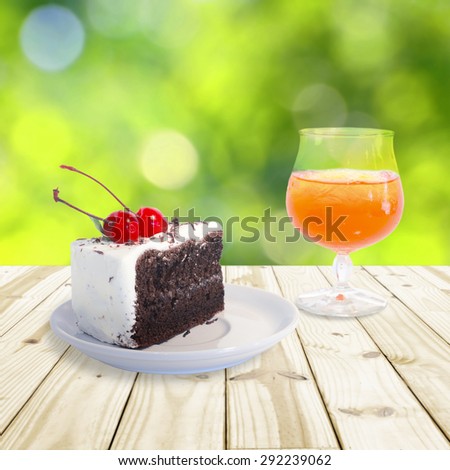 Cherry chocolate cake and Orange juice setting on wood table.  Green blurry background.  Soft focus on cherry