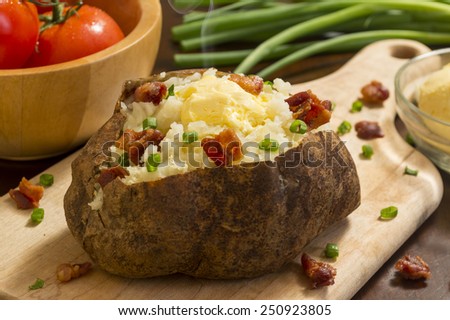 hot baked potato topped with bacon and chives on a cutting board with vegetables behind\