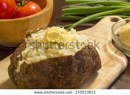hot baked potato with butter on a cutting board with vegetables behind