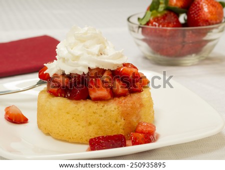 Bright red strawberries topped with syrup and whipped cream on a yellow shortcake. Served on a square plate set on a white table cloth. Bowl of strawberries in the background.\