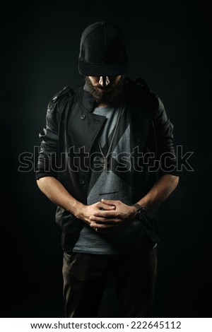 The brutal bearded man in a black jacket stands with his head bowed and holding hands clasped