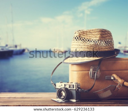Vintage suitcase, hipster hat, photo camera and passport on wooden deck. Tropical sea, beach and yachts in background. Summer holiday  and cruise traveling concept.
