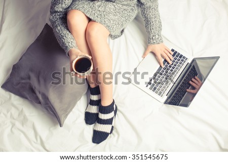 Color picture of a beautiful young woman drinking coffee at home in her bed wearing a cozy sweater while checking her laptop