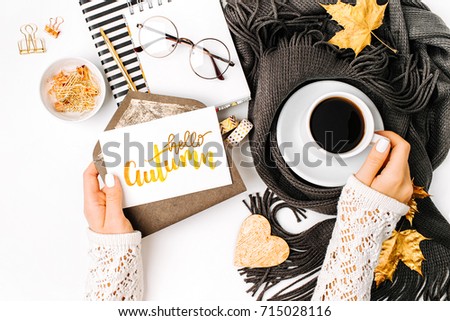 Woman holding card with word Hello Autumn. Workspace with laptop,  coffee cup wrapped in scarf,   glasses. Stylish office desk. Autumn  concept.  Flat lay, top view