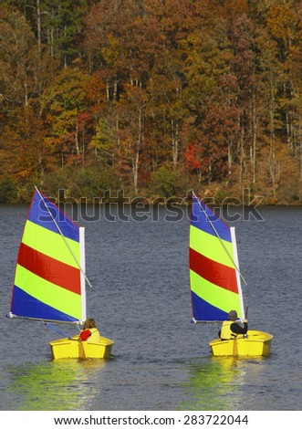 Chilly Beginnings: Two young people learning to sail in small boats on a lake with fall foliage in the background.