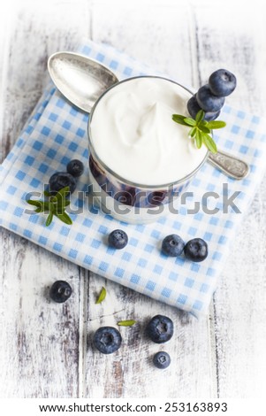 Light breakfast setup with yogurt. Plane Greek style yogurt with jam and loose blueberries on blue checkered cloth over white painted grunge style wooden table.