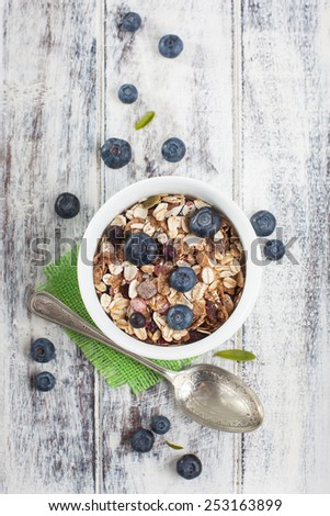 Light breakfast setup with bowl of muesli, metal spoon and fresh blueberries on green square coaster over white painted grunge style wooden table