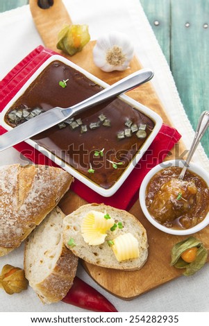 White pate dish with knife, bread, butter, chutney, tomatoes, peppers and garlic on red cloth over wooden cutting board