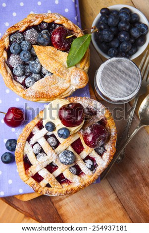 Decorated homemade shortcrust pastry berry pies with polka dot cloth, shiny metal icing sugar shaker, spoon, fork and selection of berries on grunge style wooden table