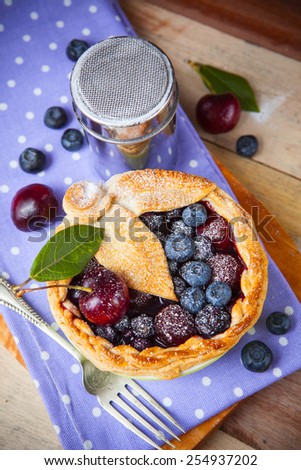 Decorated homemade shortcrust pastry berry pie with polka dot cloth, shiny metal icing sugar shaker, fork and selection of berries on grunge style wooden table.