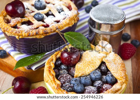 Two decorated homemade shortcrust pastry berry pies with striped cloth, shiny metal icing sugar shaker and selection of berries on grunge style wooden table.