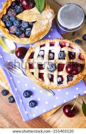 Two decorated homemade shortcrust pastry berry pies with polka dot cloth, shiny metal icing sugar shaker, fork, spoon and selection of berries on grunge style wooden table.