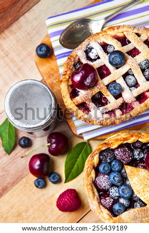 Two decorated homemade shortcrust pastry berry pies with striped cloth, shiny metal icing sugar shaker, spoon and selection of berries on grunge style wooden table.