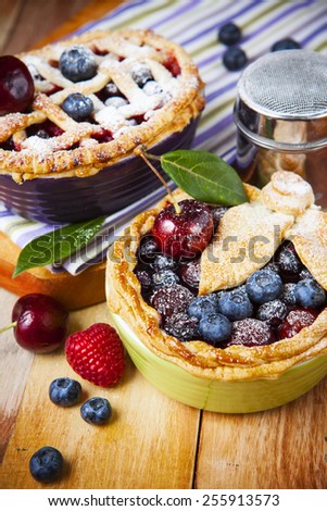 Two decorated homemade shortcrust pastry berry pies with striped cloth, shiny metal icing sugar shaker and selection of berries on grunge style wooden table.