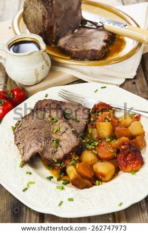 Slices of roast beef and garnish with fork on vintage plate over rustic wooden table with tomatoes, jug of sauce, roast beef joint on oval plate and carving set on the background