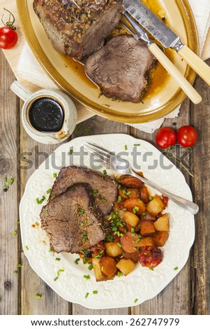 Slices of roast beef garnished with vegetables served on vintage plate and jug of sauce on rustic wooden table with roast beef joint on oval plate and carving set