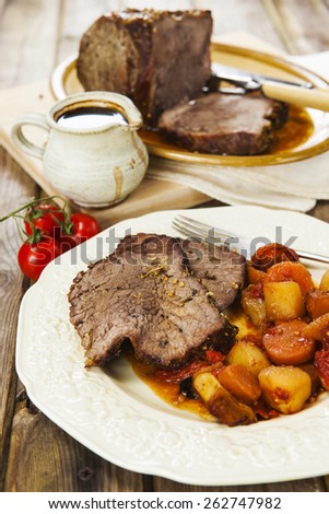 Slices of roast beef and garnish with fork on vintage plate over rustic wooden table with tomatoes, jug of sauce, roast beef joint on oval plate and carving set on the background
