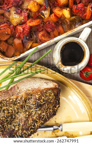 Roast beef joint with carving knife and fork on oval plate over rustic wooden table with pan of roasted vegetables and jug of sauce