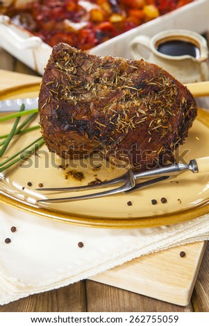 Roast beef joint with chives, carving knife and fork on oval plate over rustic wooden table with pan of roasted vegetables and jug of sauce on the background