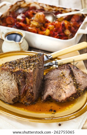 Carved roast beef joint with carving knife and fork on oval plate over rustic wooden table with pan of roasted vegetables and jug of sauce on the background