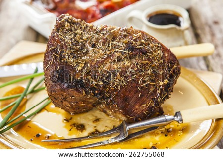 Roast beef joint with chives, carving knife and fork on oval plate over rustic wooden table with pan of roasted vegetables and jug of sauce on the background