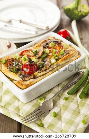 Quiche Lorraine served in white baking dish on checkered potholder with fork, plates, spring onions, broccoli and tomato over rustic wooden table