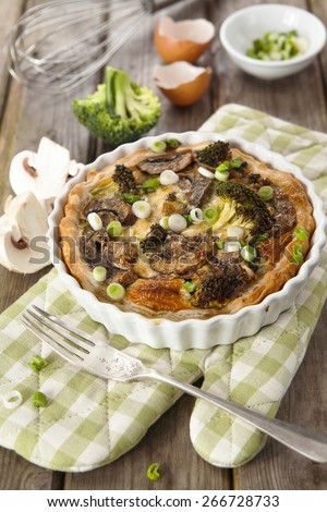 Quiche Lorraine served in round white baking dish on checkered oven-glove with mushrooms, spring onions and fork over rustic wooden table