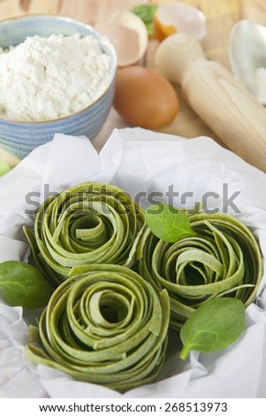 Raw homemade spinach pasta nests with spinach leaves, eggs, bowl of flour and rolling pin over the rustic wooden table