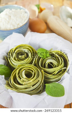 Raw homemade spinach pasta nests with spinach leaves, eggs, bowl of flour and rolling pin over the rustic wooden table