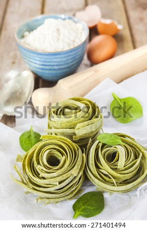 Raw homemade spinach pasta nests with spinach leaves, eggs, bowl of flour, spoon and rolling pin over the rustic wooden table