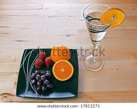 A cocktail drink in fancy glass and frosted rim with orange slice, and a plate with strawberries grapes and oranges.